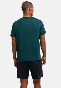 Mężczyzna z krótkimi włosami, ubrany w ciemnozieloną koszulkę z krótkim rękawem i czarne spodenki, stoi tyłem do widza.