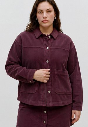 Femme portant une veste en velours côtelé violet foncé et une jupe assortie avec des boutons-pression argentés, debout devant un fond clair uni.