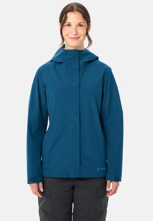 Frau trägt eine blaue wasserfeste Kapuzenjacke und schwarze Cargohosen, steht und lächelt vor einem einfarbigen Hintergrund.