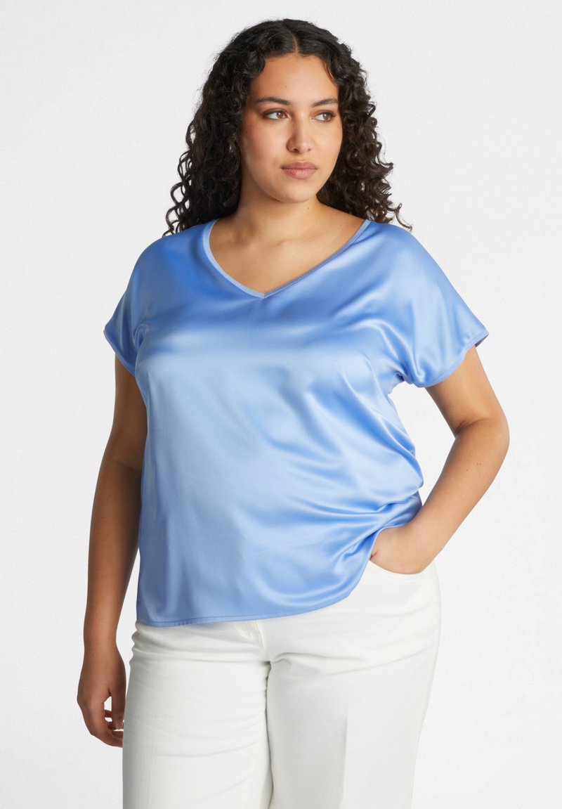 Vaaleansininen satiinipaita, jossa on V-pääntie, lyhyet hihat ja rento malli, päällä valkoiset housut. Paidassa on sileä pinta ja hienovarainen kiilto.