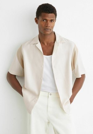 Mees, kellel on lühikesed lokkis juuksed ja kes kannab beežikat lühikeste varrukatega nööbitavat särki valge tankitopi ja valgete pükstega, seisab käed taskutes.