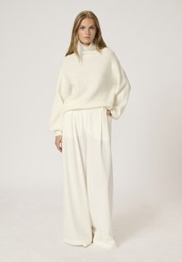 Pull en tricot oversize de couleur blanc crémeux avec un col montant et des manches ballon, associé à un pantalon large crème avec des poches latérales.