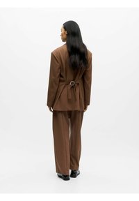 Bruine blazer en broek met wijde pijpen, op maat gemaakt ontwerp met een riemdetail aan de achterkant, gladde textuur en glanzende zwarte schoenen.