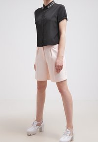 Zwarte cropped top met versierde kraag, gecombineerd met lichtroze shorts. Model draagt witte block-heeled schoenen met vetersluiting.