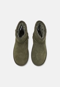 Botas de tobillo de gamuza verde con un interior suave, forma redondeada, detalle de correa lateral y suela texturizada. Diseñadas para brindar calor y comodidad.