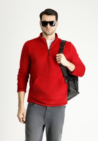 Suéter de polar rojo con diseño de media cremallera, bolsillo delantero y puños de canalé. Combinado con jeans grises y un bolso negro de hombro.