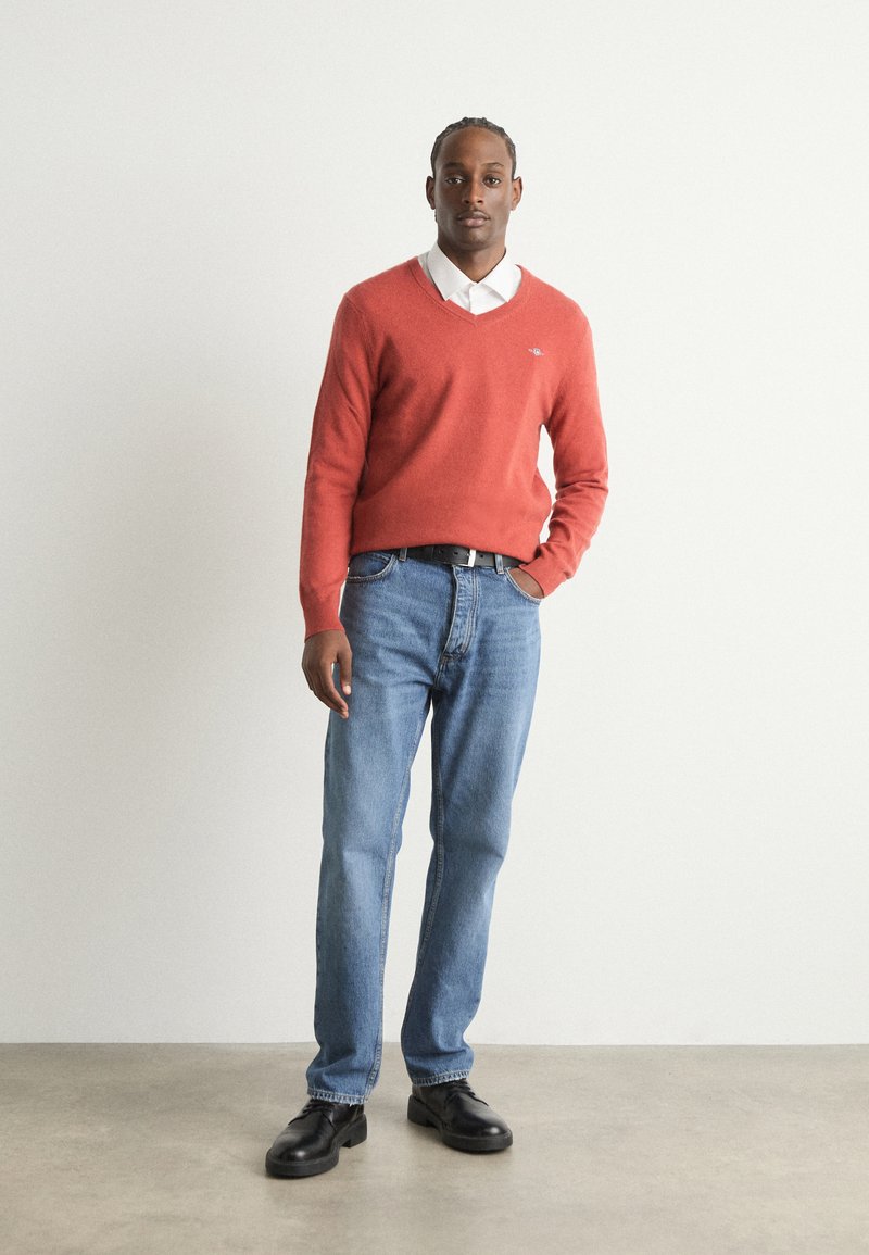 Roter V-Ausschnitt-Pullover über einem weißen Hemd mit Kragen, kombiniert mit blauen Jeans und schwarzen Schuhen. Der Pullover hat ein kleines Logo auf der Brust.