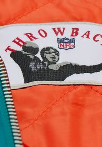 NFL Throwback-Patch auf einer gesteppten orangen Jacke, mit einem schwarz-weißen Grafikdesign, das Spieler und Text zeigt, umrandet von einem hellen Saum.
