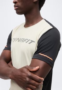 Άνδρας με σταυρωμένα χέρια, που φοράει μια αθλητική μπλούζα Dynafit μπεζ και μαύρου χρώματος με πορτοκαλί λεπτομέρειες στα μανίκια, μπροστά σε απλό φόντο.