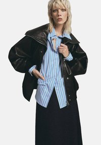 Chaqueta oversized de cuero negro sobre una camisa de rayas azul claro y blanca, combinada con una falda oscura. Materiales texturizados y contrastes audaces.