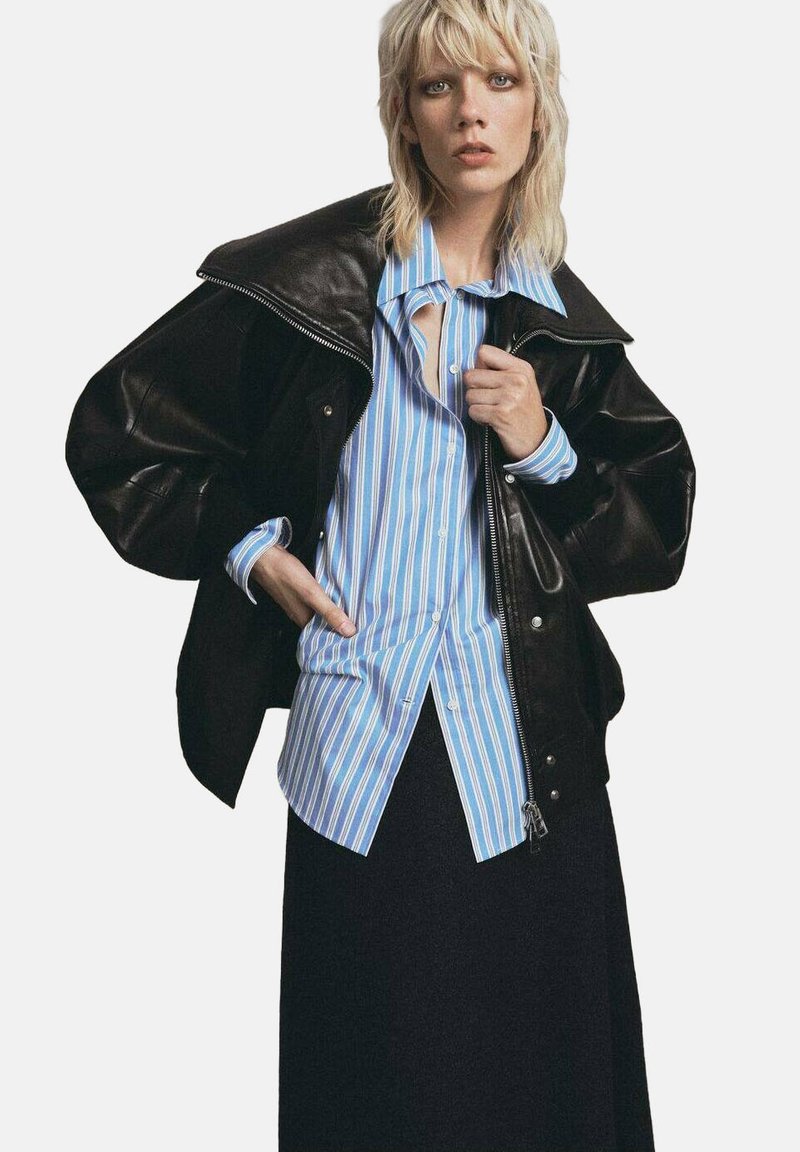 Chaqueta oversized de cuero negro sobre una camisa de rayas azul claro y blanca, combinada con una falda oscura. Materiales texturizados y contrastes audaces.