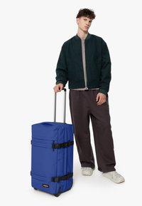 Modrý kufr na kolečkách vyrobený z odolné tkaniny, s několika popruhy a teleskopickou rukojetí, vedle osoby v tmavě zelené bundě a šedých kalhotách.