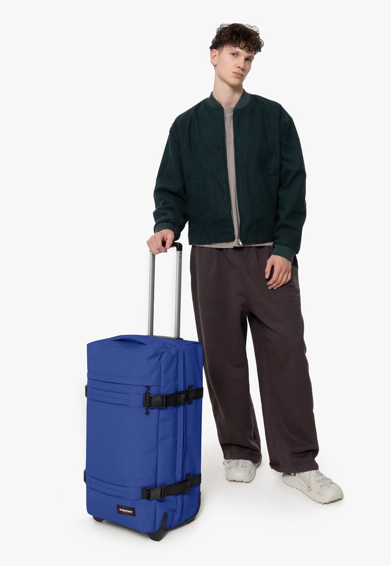 Modrý kufr na kolečkách vyrobený z odolné tkaniny, s několika popruhy a teleskopickou rukojetí, vedle osoby v tmavě zelené bundě a šedých kalhotách.