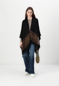 Poncho nero con bordo a stampa leopardata e strisce marroni, realizzato in tessuto morbido. Indossato sopra un top beige, abbinato a jeans blu e scarpe bianche.