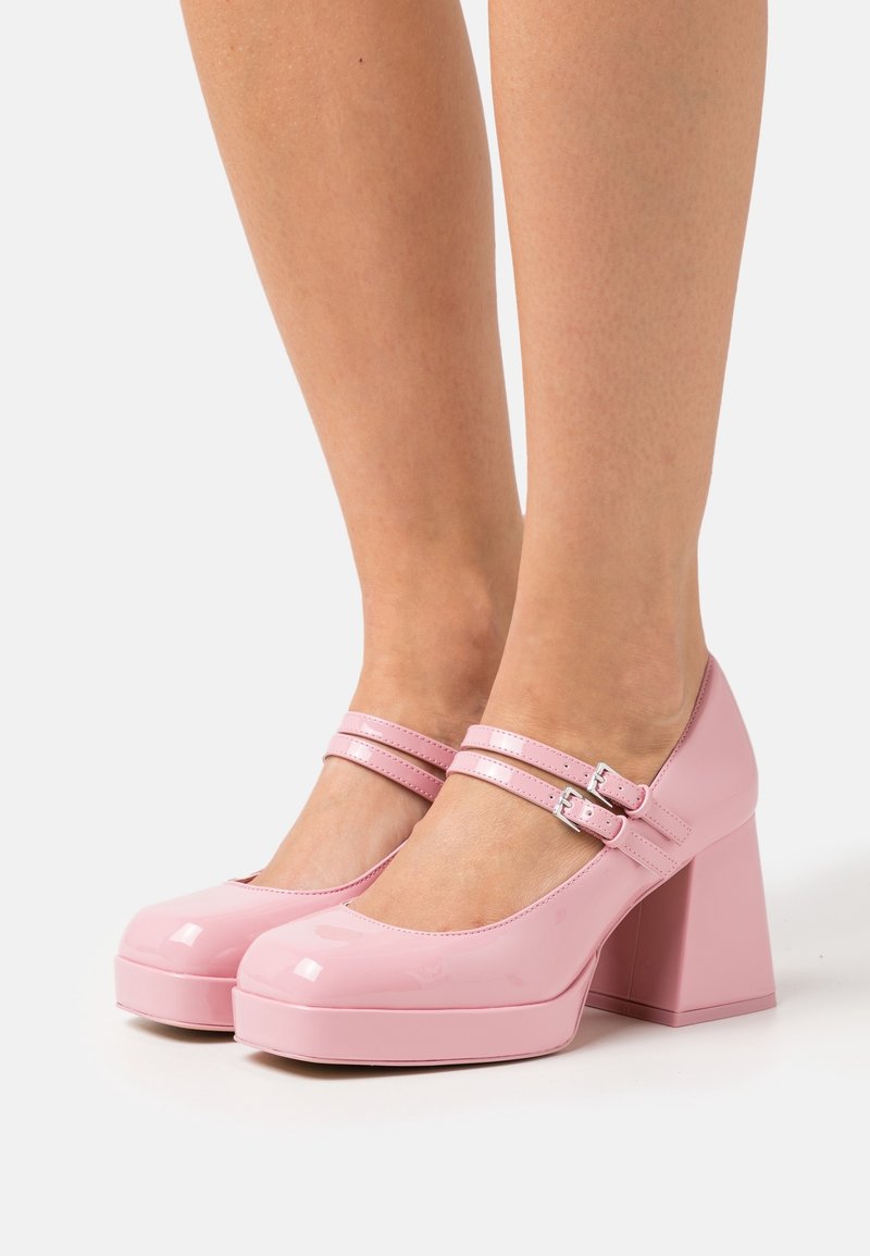 Jambes portant des talons compensés Mary Jane roses brillants avec double sangle et boucles argentées sur fond blanc.