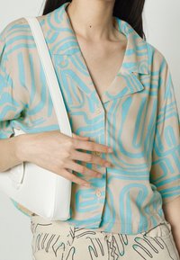 Beżowa bluzka z krótkim rękawem, z niebieskimi abstrakcyjnymi liniami, z przypinanym przodem i kołnierzykiem z wcięciem, w połączeniu z białą torebką na ramię.