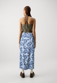 Ärmlös, olivgrön texturerad topp med justerbar knytning. Nedanför når en lång blå- och vitmönstrad kjol halvvägs ner till vaden, parat med bruna skor.