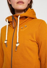 Felpa arancione con zip frontale, cordini bianchi e dettagli metallici. Presenta testo ricamato e due bottoni di legno. Texture morbida.