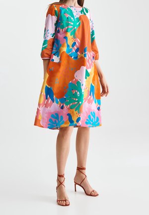 Frau trägt ein farbenfrohes, locker sitzendes Kleid mit abstrakten Blumenmustern und hautfarbene, hochhackige Sandalen mit Knöchelriemen.