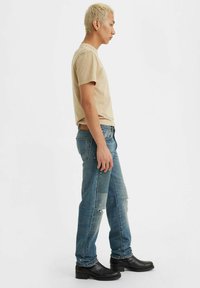 Camiseta de manga corta beige, jeans desgastados de color azul claro con apliques, tachuelas plateadas y botas negras hasta el tobillo. Modelo visto de perfil.