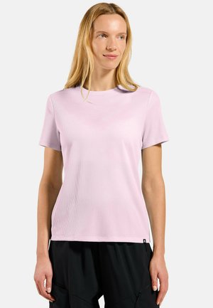 Woman with long blonde hair wearing a light pink short-sleeve textured shirt and black pants, standing against a plain light background.