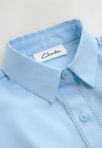Hellblaues Hemd mit einem klassischen Kragen, Knopfleiste und zartem Spitzenbesatz entlang der Knopfleiste. Mit einem "Clarks"-Label im Halsausschnitt.