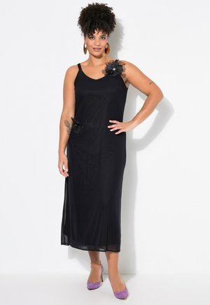 Schwarzes ärmelloses Kleid mit rundem Ausschnitt, verziert mit Lace-Blumenakzenten an der Schulter und Hüfte, kombiniert mit lila Absatzschuhen.