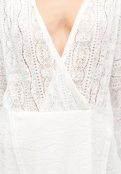 Λευκό φόρεμα από δαντέλα με βαθύ V-λαιμόκοψη, μακριά μανίκια και υφασμάτινα σχέδια. Η δαντέλα διαθέτει περίπλοκα μοτίβα και ομαλή βάση υφάσματος.