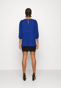 Top blu, a maniche lunghe, con apertura a chiave sul retro, indossato sopra una mini gonna nera. Il tessuto sembra leggero e ha una vestibilità comoda.