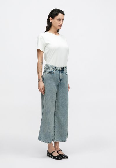 Λευκό κοντομάνικο μπλουζάκι σε συνδυασμό με τζιν παντελόνι ψηλόμεσο, φαρδύ και ανοιχτού μπλε χρώματος. Μαύρα πλατιά παπούτσια με διακοσμητικές λεπτομέρειες.