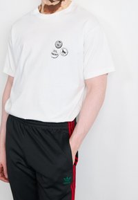 Vit bomullst-shirt med tre tryckta cirkelformade logotyper på bröstet, kombinerad med svarta träningsbyxor med röda ränder och grön varumärkeslogotyp.