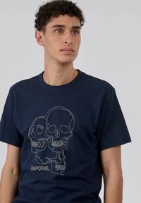 T-shirt bleu marine à manches courtes avec un design en contour blanc de deux têtes de mort et le logo "KAPORAL" en bas.