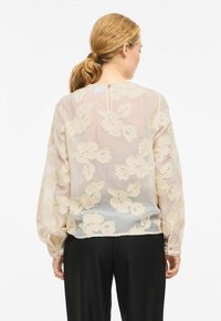 Blouse légère et transparente en beige doux, ornée de broderies florales texturées, avec manches longues bouffantes et fermeture arrière à goutte.