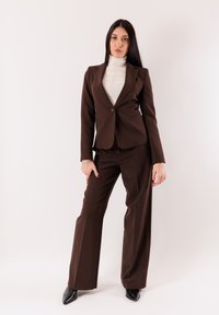 Abito marrone su misura con blazer attillato e pantaloni a gamba larga. Sotto, un dolcevita bianco a costine. Scarpe nere con punta appuntita. Sfondo semplice.