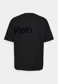 Camiseta de algodón negra con mangas cortas, que presenta la palabra "Klein" en un estampado negro brillante en la parte posterior. Diseño de cuello redondo, ajuste relajado.