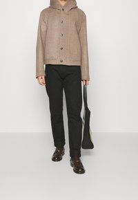 Person som bär en brun huva jacka, svarta byxor och mörkbruna skor, och håller en svart tygväska med neongröna detaljer.