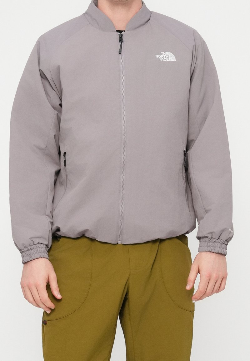 Homme portant une veste zippée gris clair avec des poignets élastiques et un pantalon vert, debout devant un fond blanc uni.