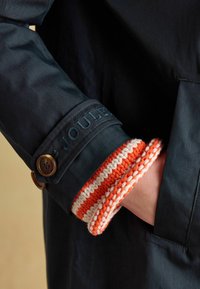 Ręka osoby w kieszeni płaszcza pokazująca ciemnogranatowy rękaw z brązowymi guzikami oraz pod spodem pomarańczowo-biały prążkowany, dziergany mankiet.