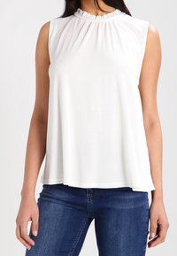 Valkoinen hihaton toppi, jossa on röyhelökaulus, rento malli ja sileä pinta, yhdistettynä sinisiin farkkuihin.