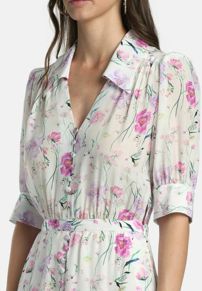 Femme portant une robe blanche à fleurs roses et violettes, manches bouffantes, boutons recouverts de tissu et taille ceinturée.