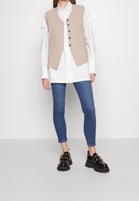 Chaleco sin mangas beige con cinco botones oscuros, combinado con una camisa blanca y jeans ajustados azules. Los zapatos negros tienen detalles dorados.