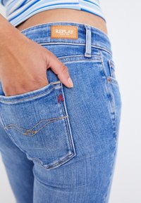 Jeans de mezclilla con un lavado azul, que presentan un diseño de bolsillo cosido, parche del logo Replay en la cinturilla y un detalle de bordado en rojo en contraste.