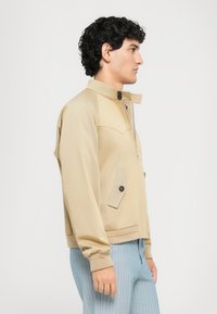 Beige lahka jakna z majhnim ovratnikom, sprednjim žepom, zadrgo in rebrastimi manšetami. Nošena preko modrih hlač z žičnimi črtami.