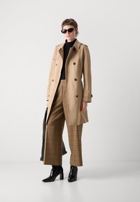 Beige trenchcoat med dubbelknäppning, svart polotröja, bruna rutiga byxor och svarta klackstövlar. Accessoarer inkluderar svarta solglasögon.