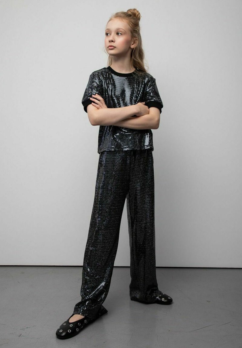 Czarny top z cekinami i pasujące szerokie spodnie o błyszczącej fakturze. Strój ma luźny krój i krótkie rękawy, uzupełniony o czarne buty wsuwane.