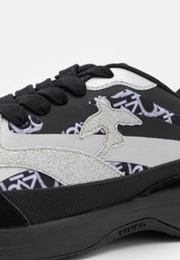 Baskets noires présentant un mélange de matériaux synthétiques lisses et d'accents de paillettes texturées, avec un design d'oiseau stylisé et des sections à motifs.