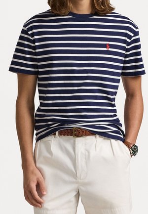 Mężczyzna w granatowo-białej poziomo paskowanej koszulce z czerwonym haftowanym logo, białych szortach, brązowym plecionym pasku i zegarku na nadgarstku.