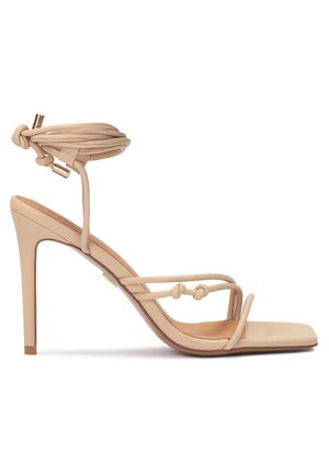 Sandale beige à talon haut avec fines brides nouées, bout carré et liens enroulés autour de la cheville avec embouts métalliques dorés.