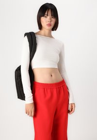 Un crop top blanc à manches longues associé à un pantalon ample rouge vif. Un sac à dos noir rembourré est porté sur une épaule.