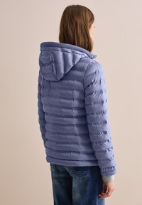 Ljuslila pufferjacka med en strukturerad, glänsande yta, med huva och horisontell quiltning. Matchad med blå jeans.
